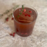 Sparkling pomegranate mocktail served over ice in a short glass, garnished with fresh thyme and pomegranate arils on a stone background