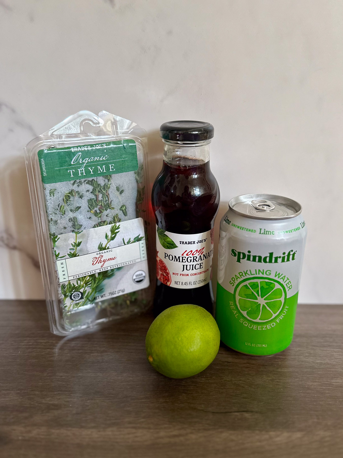 Ingredients for a sparkling pomegranate mocktail, including Trader Joe’s organic thyme, 100% pomegranate juice (not from concentrate), a fresh lime, and Spindrift lime sparkling water.
