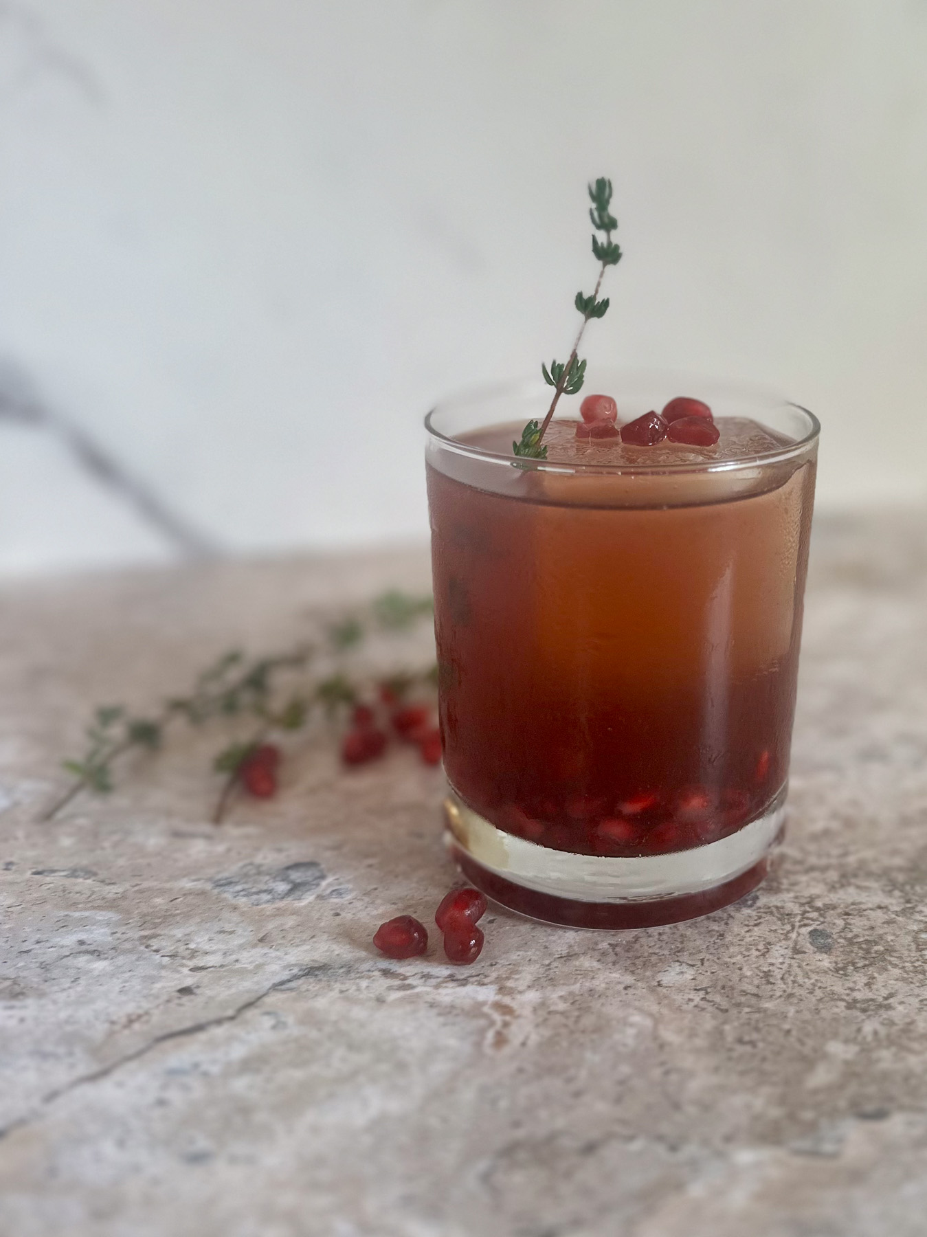 Sparkling pomegranate mocktail served in a short glass over ice, garnished with a thyme sprig and pomegranate arils, with fresh thyme and arils scattered on a stone countertop.