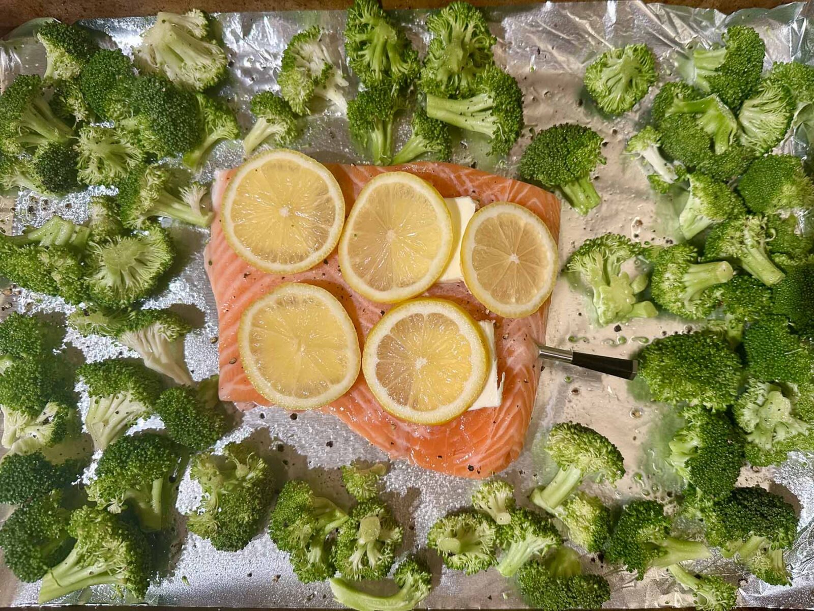 Uncooked sheet pan meal with salmon topped with lemon slices and butter, surrounded by raw broccoli florets and a meat thermometer inserted.