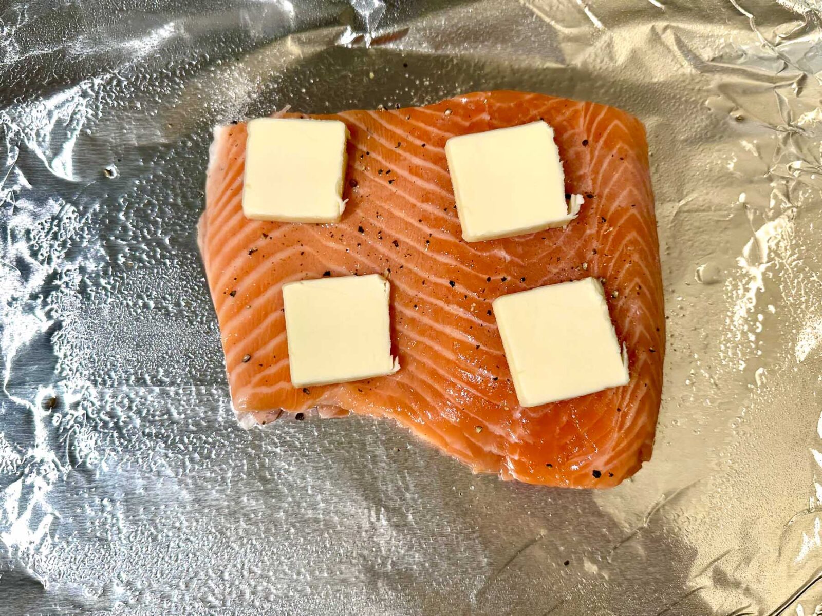 Raw salmon fillet on a foil-lined baking sheet, seasoned and topped with four pats of butter.