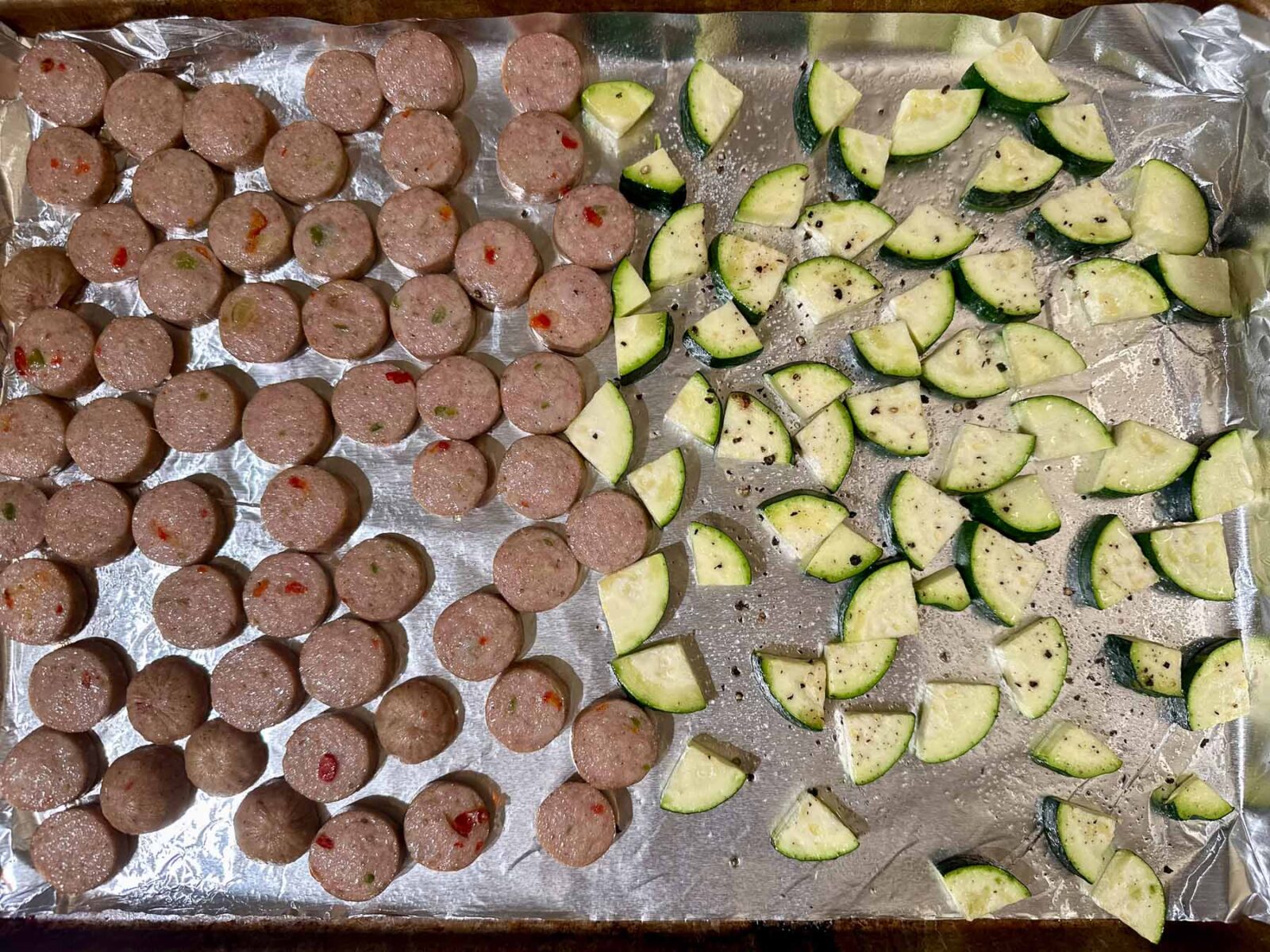 Uncooked sheet pan with sliced chicken sausage and seasoned zucchini.