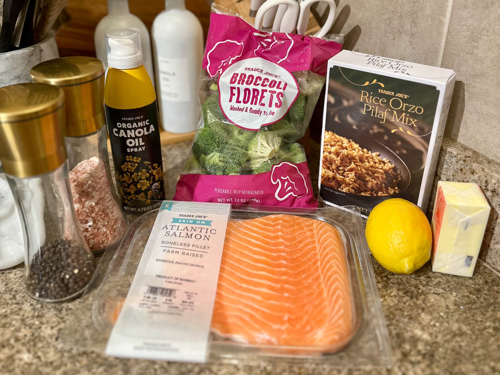 Trader Joe’s ingredients on a kitchen counter, including salmon, broccoli florets, rice orzo pilaf mix, a lemon, butter, canola oil spray, salt, and pepper.