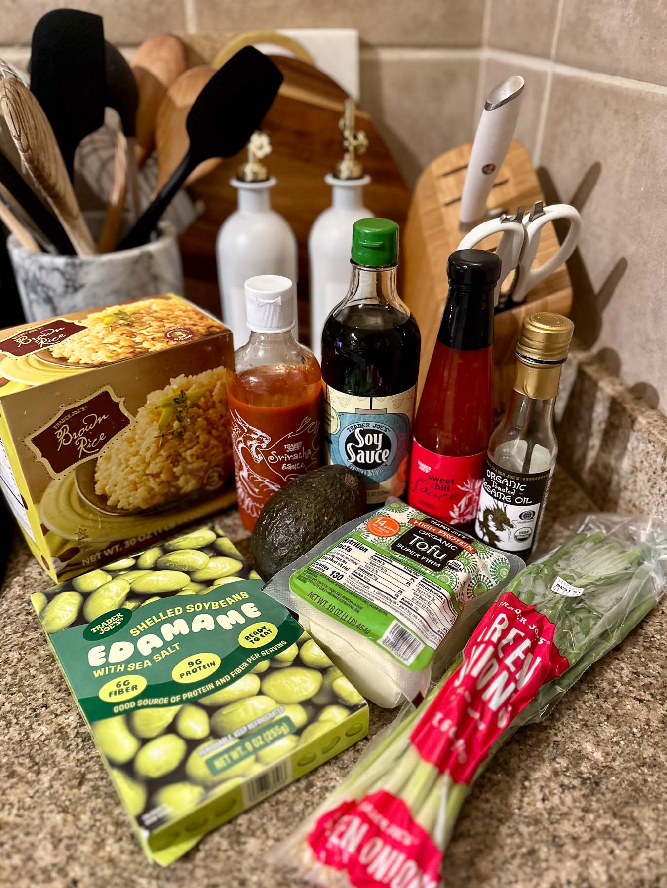 Trader Joe’s ingredients for a tofu rice bowl, including shelled edamame, tofu, avocado, green onions, brown rice, soy sauce, sriracha, sweet chili sauce, and sesame oil.