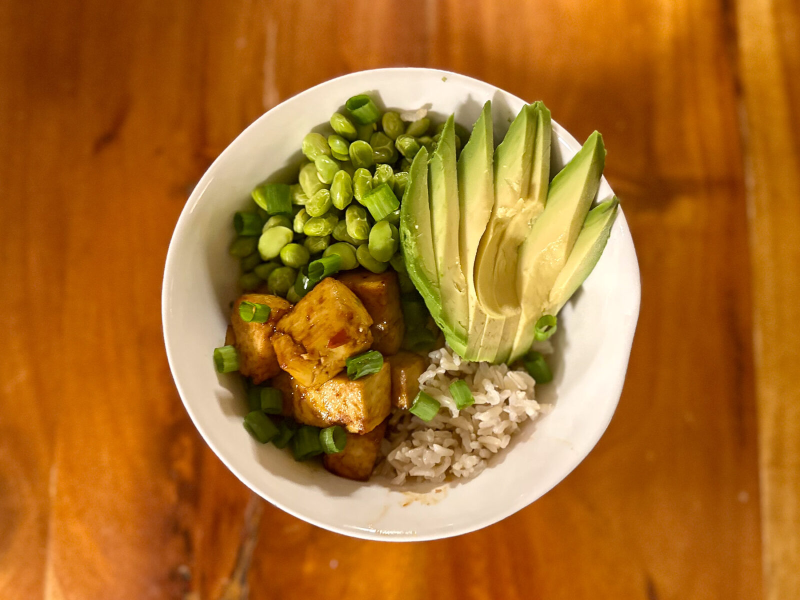 Overhead view of a vibrant tofu rice bowl with crispy tofu, edamame, sliced avocado, brown rice, and green onions.