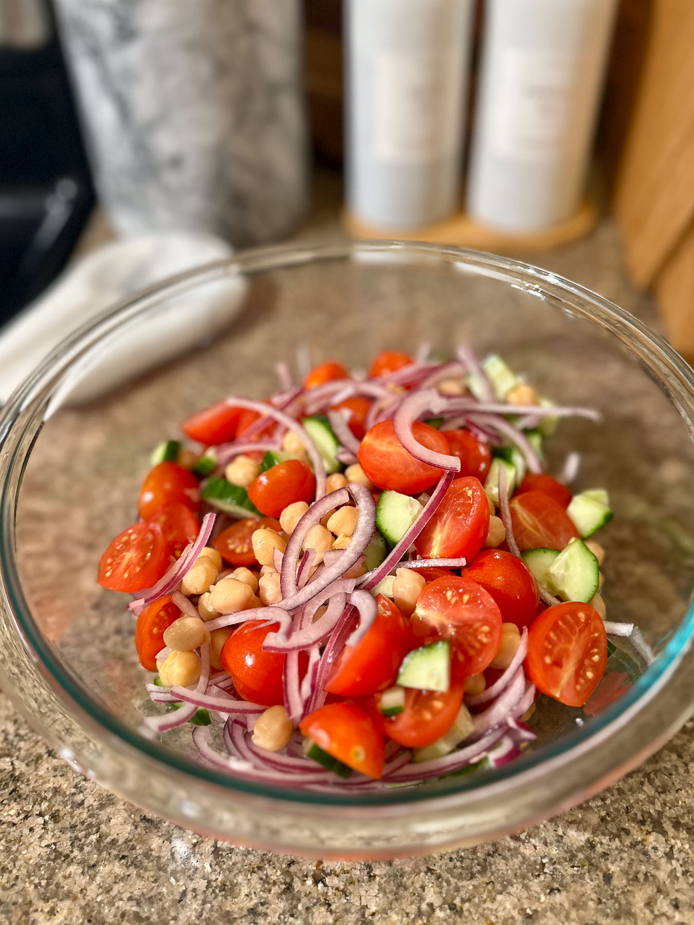 Glass bowl filled with chopped cherry tomatoes, sliced red onion, diced cucumber, and chickpeas for a Mediterranean salad base.
