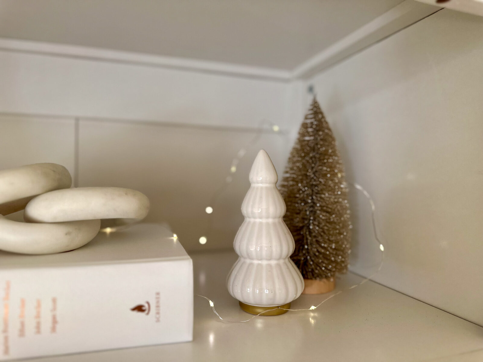 Close-up of a white ceramic tree figurine with gold trim, flanked by a sparkly bottlebrush tree and soft white fairy lights on a bookshelf.