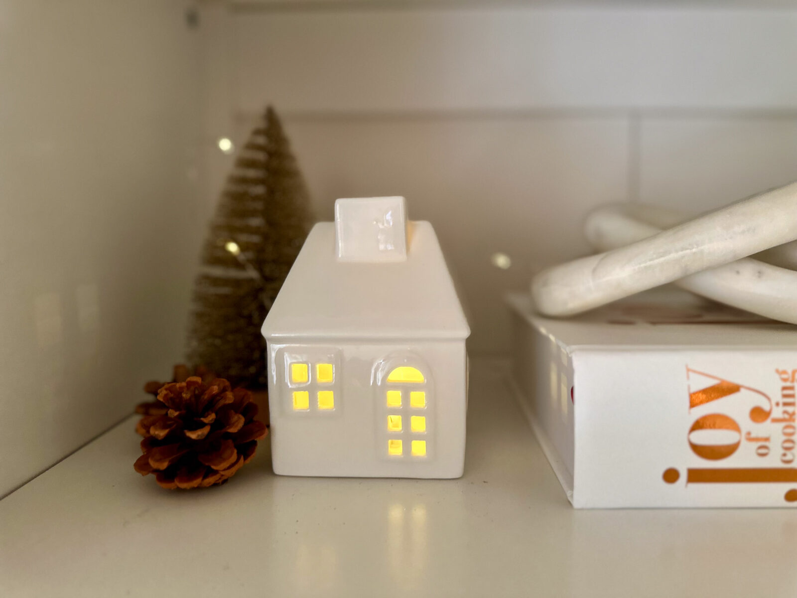 White ceramic house with glowing windows styled on a shelf with a pine cone, bottlebrush tree, and the Joy of Cooking book.