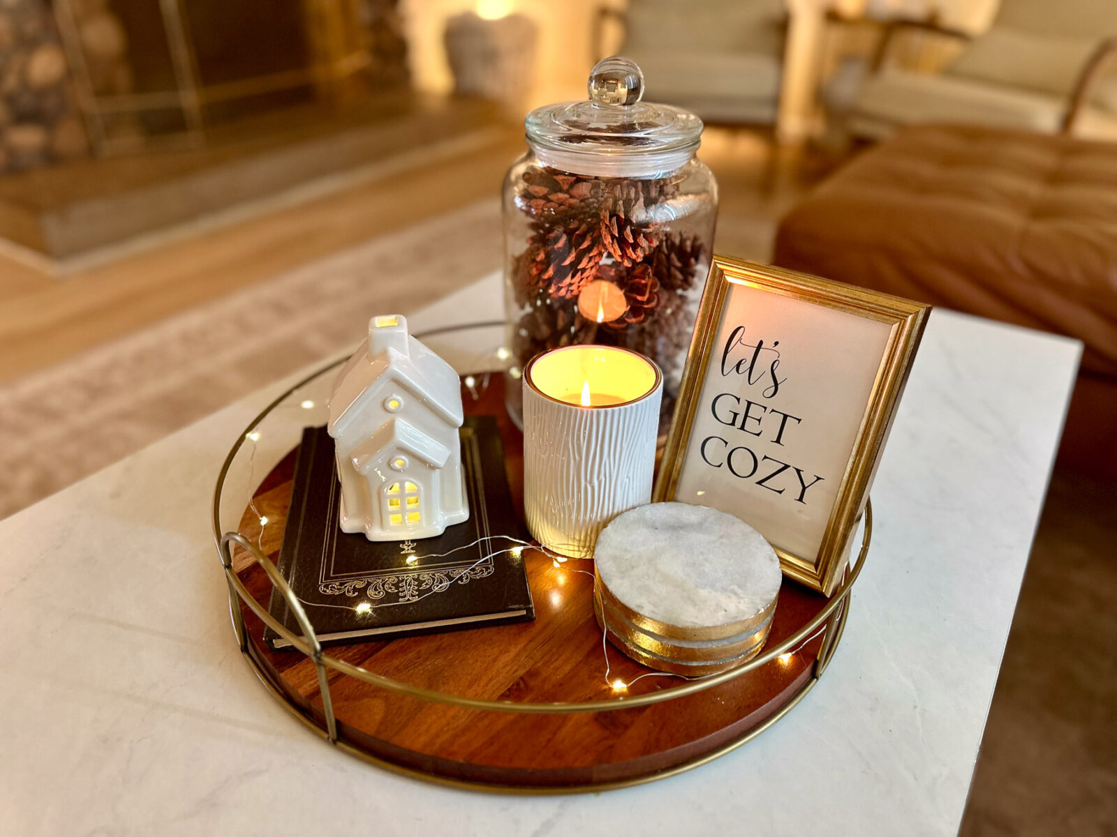 Round wood tray styled for winter with a ceramic house, lit candle, pine cone-filled jar, and framed “Let’s Get Cozy” print.