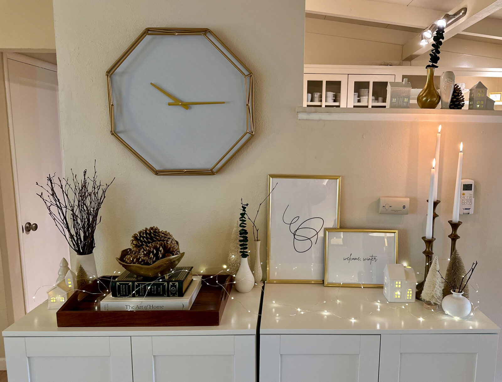 Console styling under a modern octagonal wall clock, featuring pine cones, vases, framed winter prints, and glowing ceramic houses.