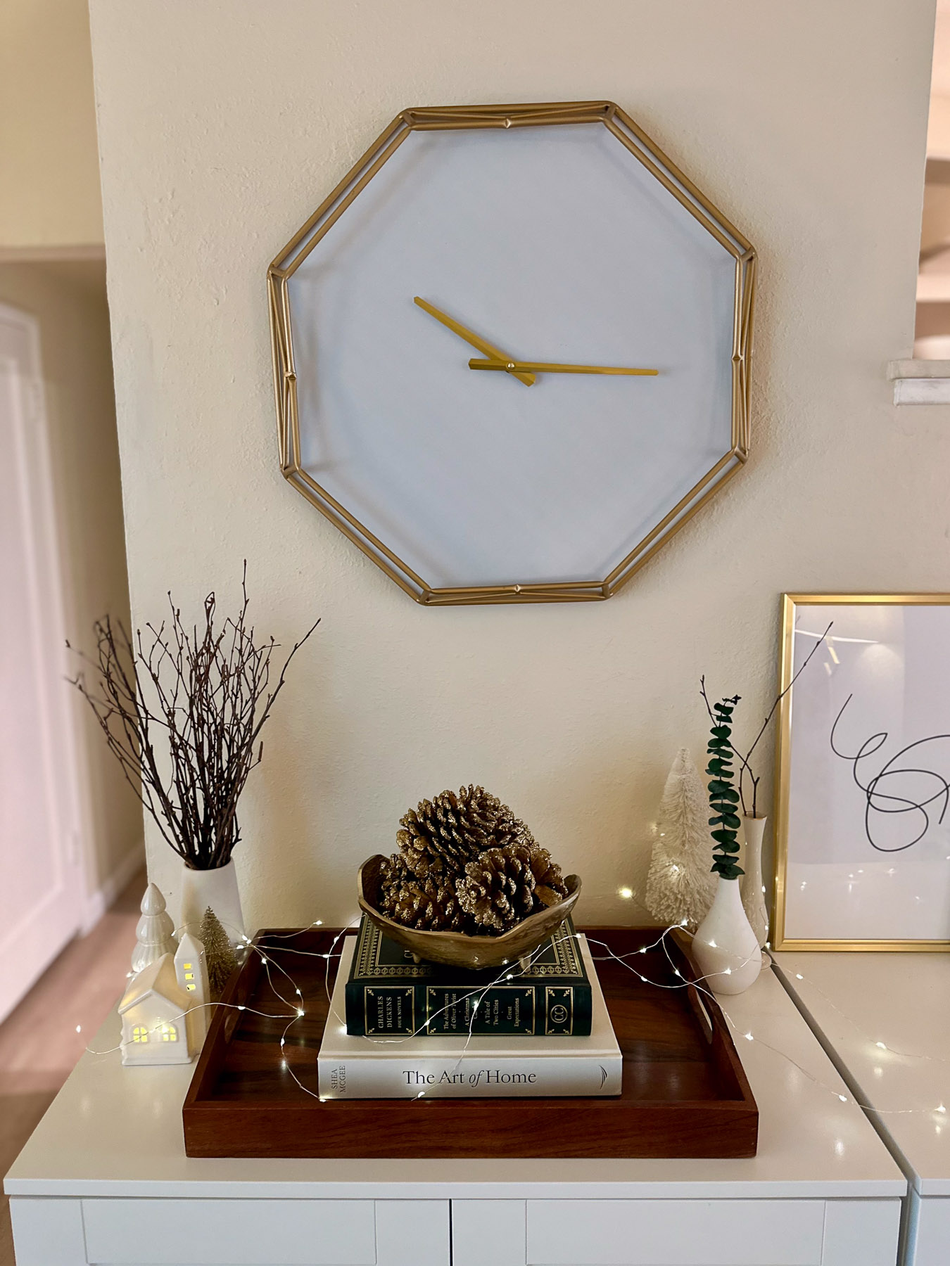 A white console table beneath a modern gold-rimmed octagon clock, styled with a tray of stacked books, ceramic houses, and pinecones. A framed line art print leans against the wall beside bare branches in a white vase.