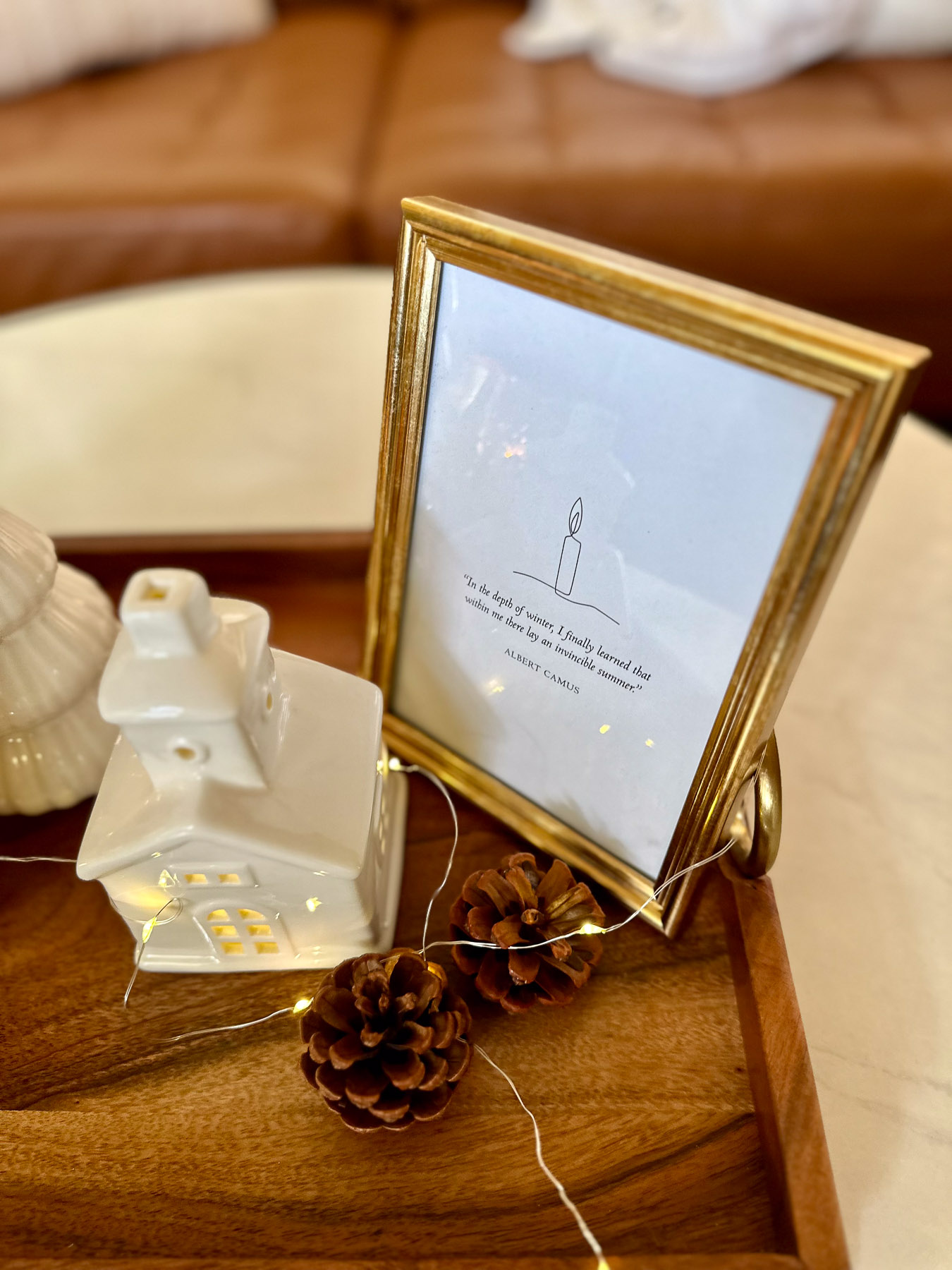 A cozy winter-themed vignette on a wooden tray featuring a gold-framed print of the Albert Camus quote “In the depth of winter, I finally learned that within me there lay an invincible summer,” illustrated with a candle drawing. A ceramic house with lit windows, a white tree figurine, and two pinecones complete the scene, with fairy lights wrapped around the tray for a soft glow.