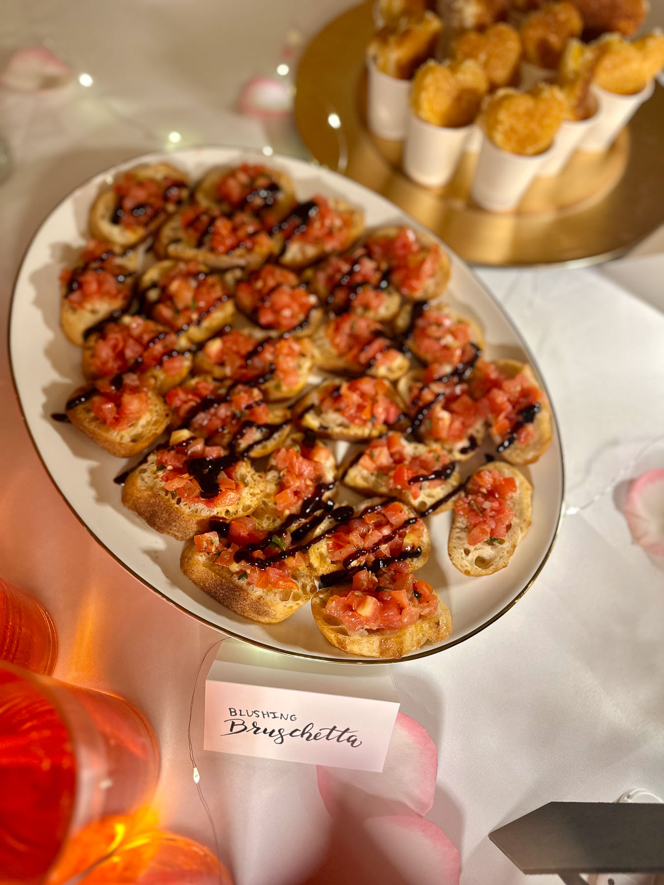 Close-up of a plate of bruschetta drizzled with balsamic glaze, labeled “Blushing Bruschetta,” served on a white party table with rose petals and twinkle lights.