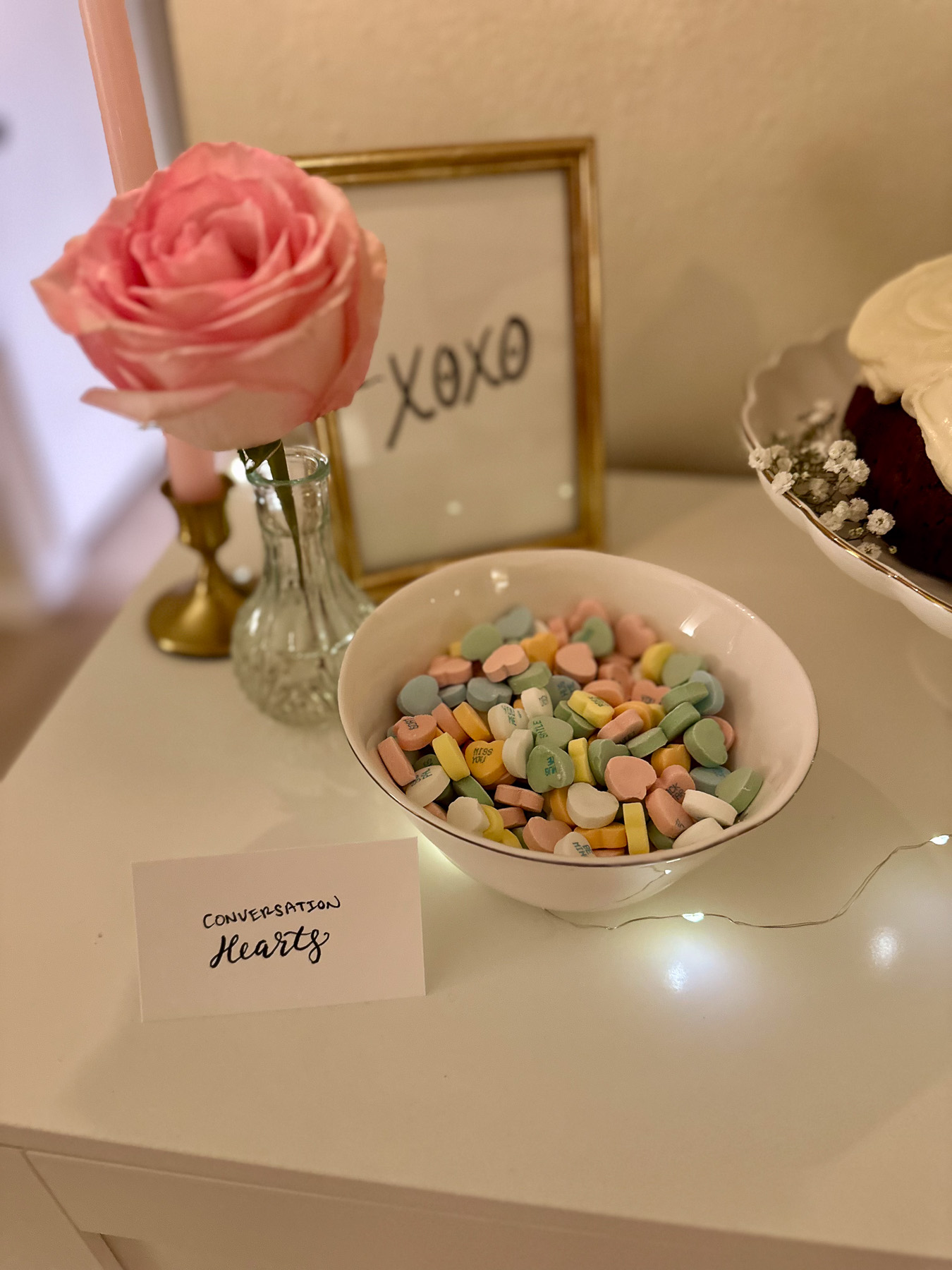 A white bowl filled with pastel-colored conversation heart candies, sitting next to a pink rose in a vase and a framed “XOXO” sign. A soft string of fairy lights runs along the tabletop.