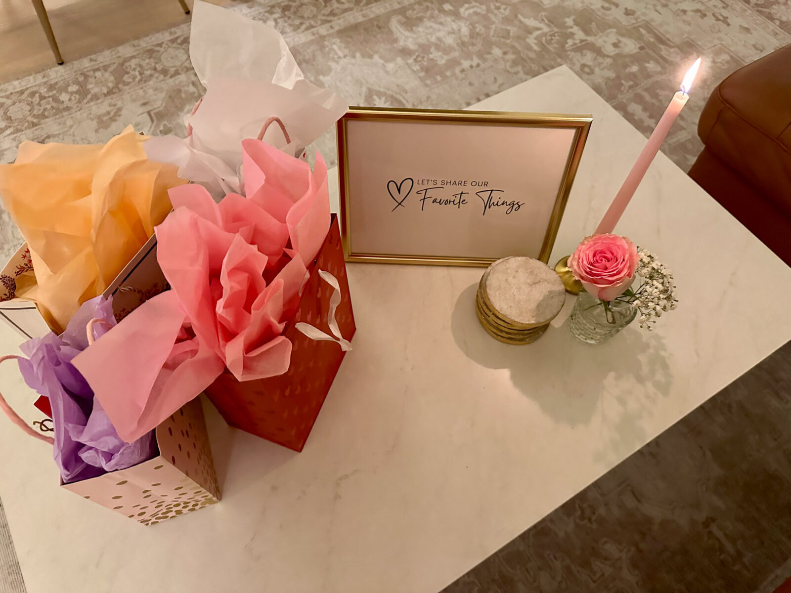A coffee table set up with pastel-colored gift bags and a framed sign that reads “Let’s Share Our Favorite Things.” A single pink taper candle and a rose in a bud vase add charm.