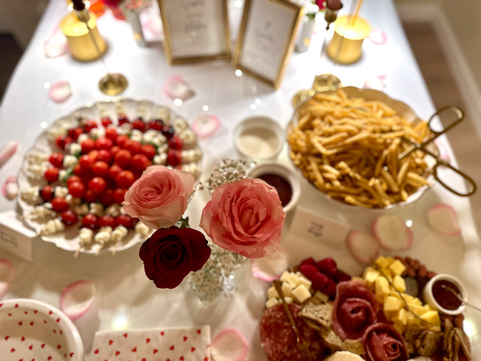 Close-up of a Galentine’s Day food table featuring a Caprese skewer platter, French fries in a white bowl, dipping sauces, and a rose centerpiece on a white tablecloth with rose petals and twinkle lights.