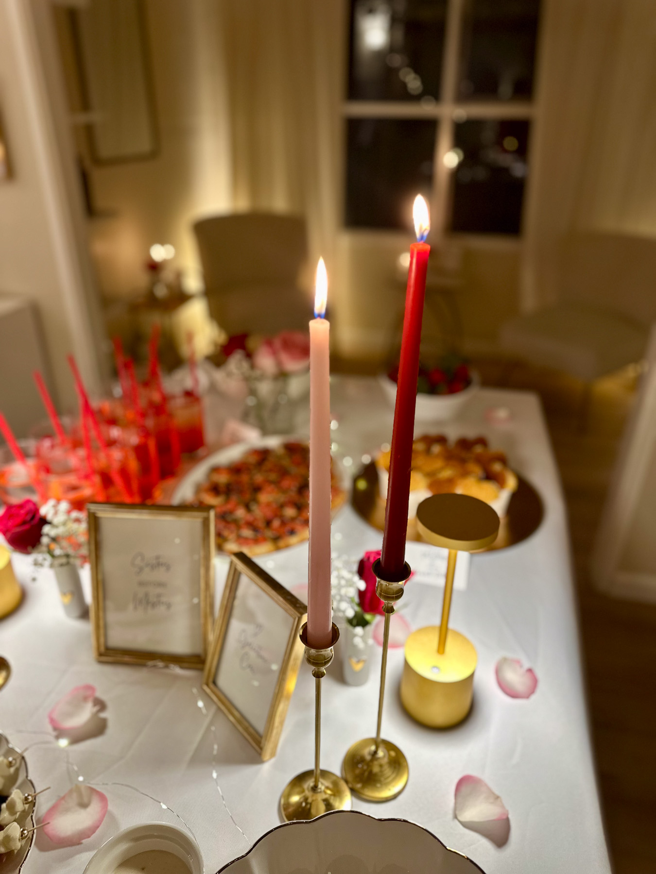 Red and pink taper candles lit on a white party table surrounded by gold candle holders, framed Galentine’s quotes, and a spread of themed food in the background.