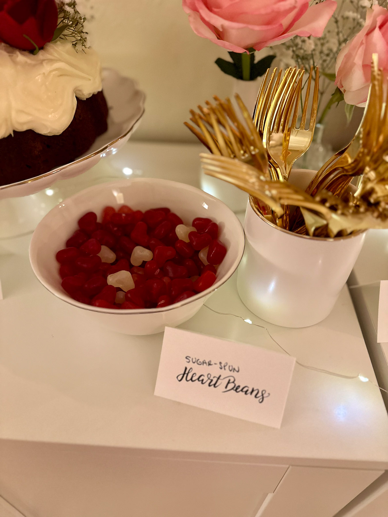 A white bowl of red and white heart-shaped jelly beans labeled “Sugar-Spun Heart Beans,” next to a white container of gold forks and a pink rose centerpiece.