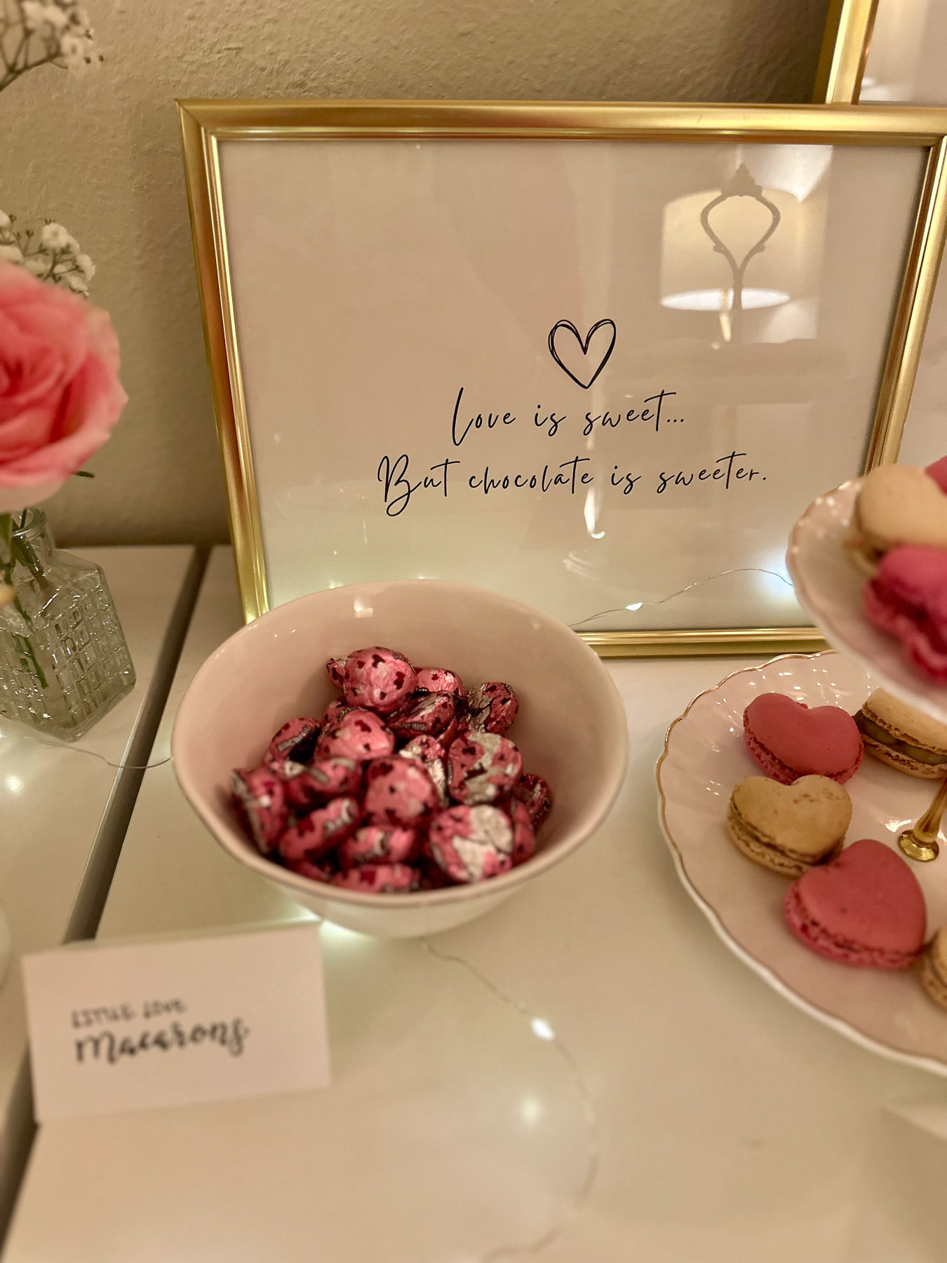 A bowl of foil-wrapped pink heart-shaped chocolates labeled beside a framed quote: “Love is sweet… but chocolate is sweeter.” Pink roses and fairy lights complete the scene.