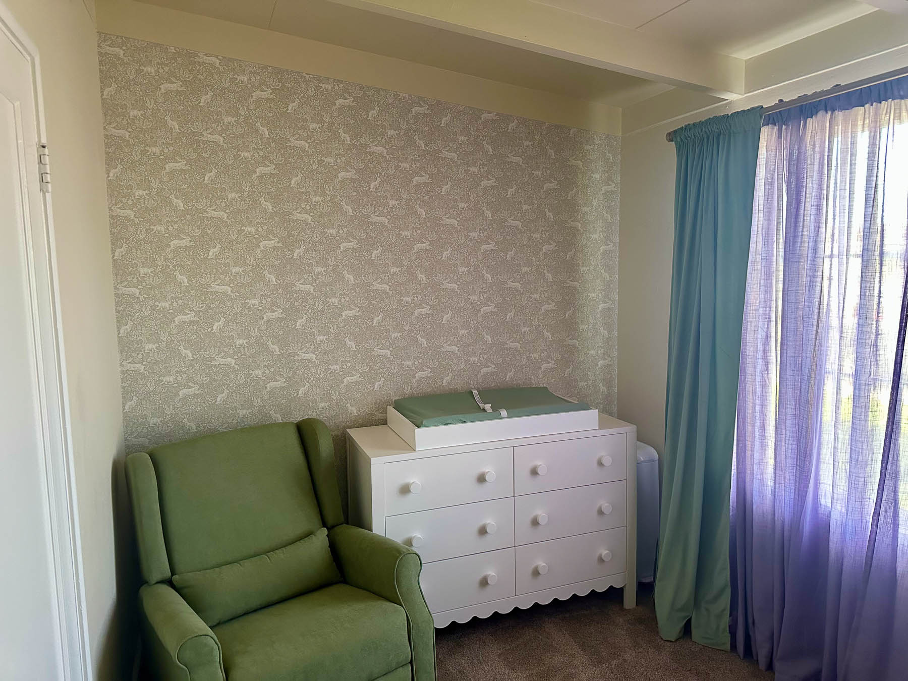 Nursery with peel-and-stick wallpaper accent wall, green armchair, white dresser, and sage and lavender curtains