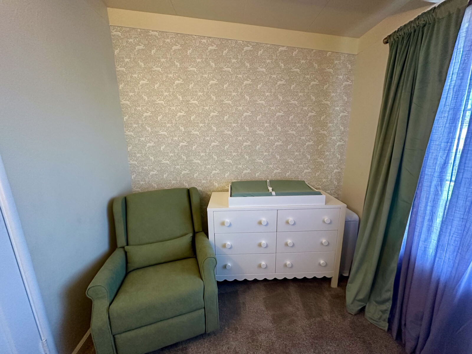 Nursery with peel-and-stick wallpaper accent wall, green armchair, white dresser, and layered green curtains.