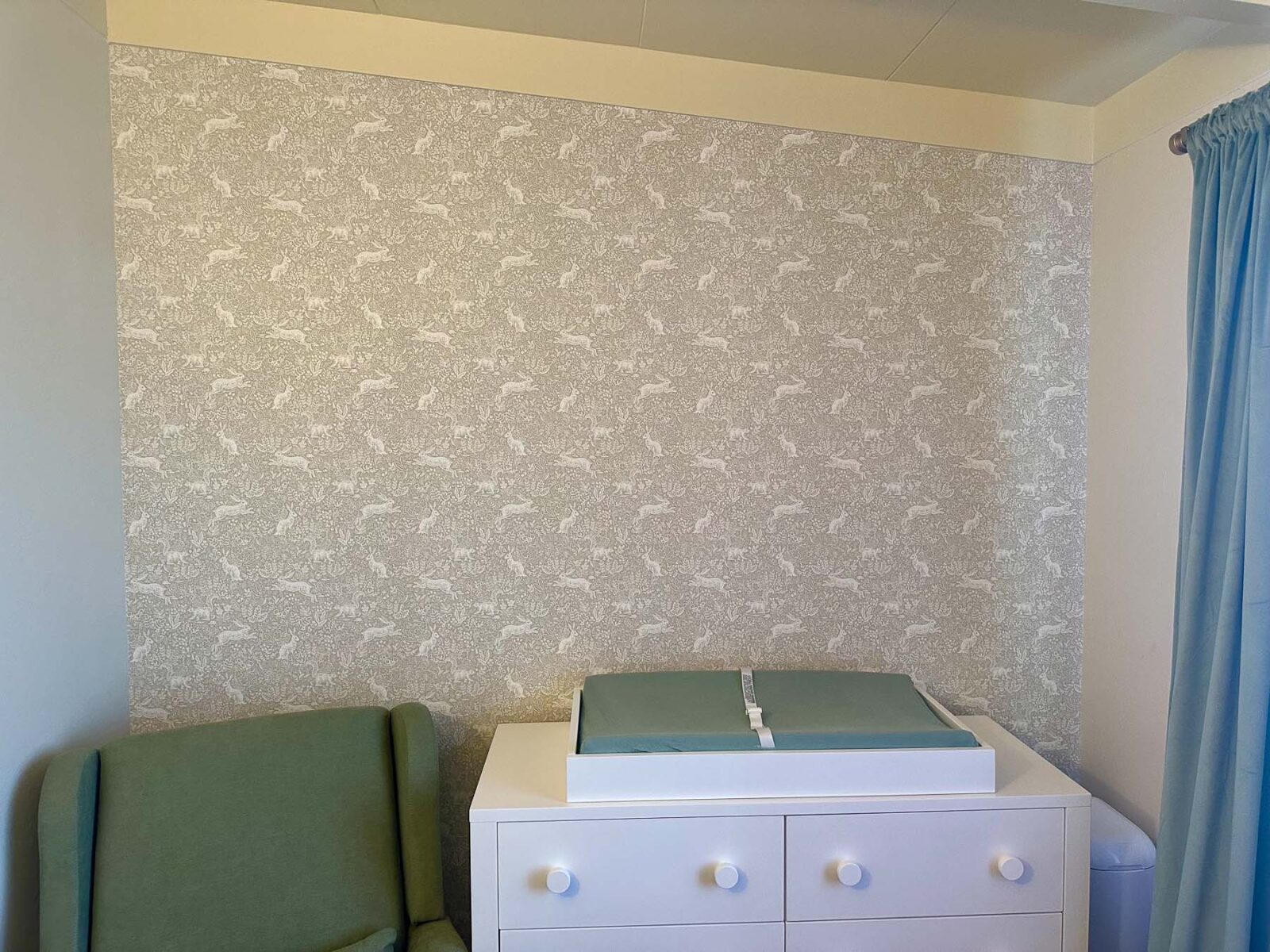 Nursery accent wall with peel-and-stick wallpaper, green chair, and white dresser.