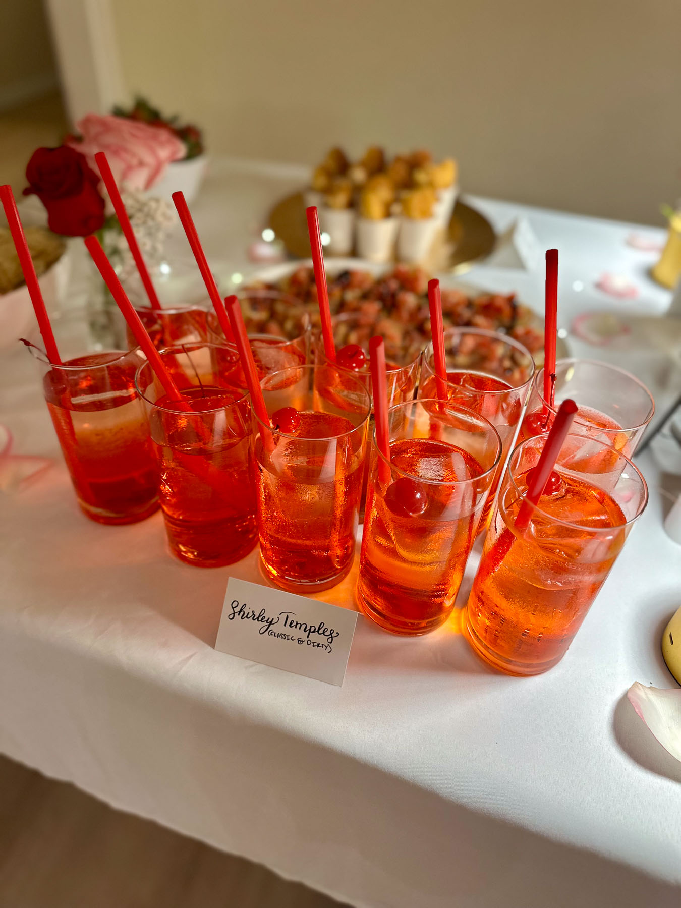 Row of bright red Shirley Temples with cherries and red straws on a Galentine’s Day table setup, with bruschetta and other appetizers in the background.