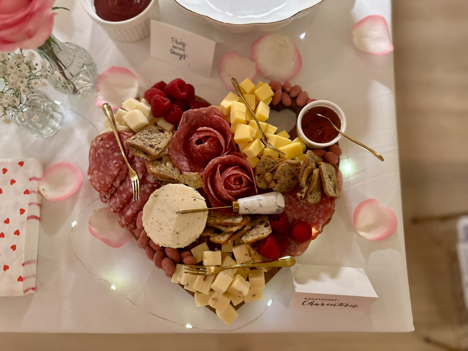 Heart-shaped charcuterie board featuring cheese, salami, raspberries, crackers, and jam, styled with gold utensils and pink rose petals.