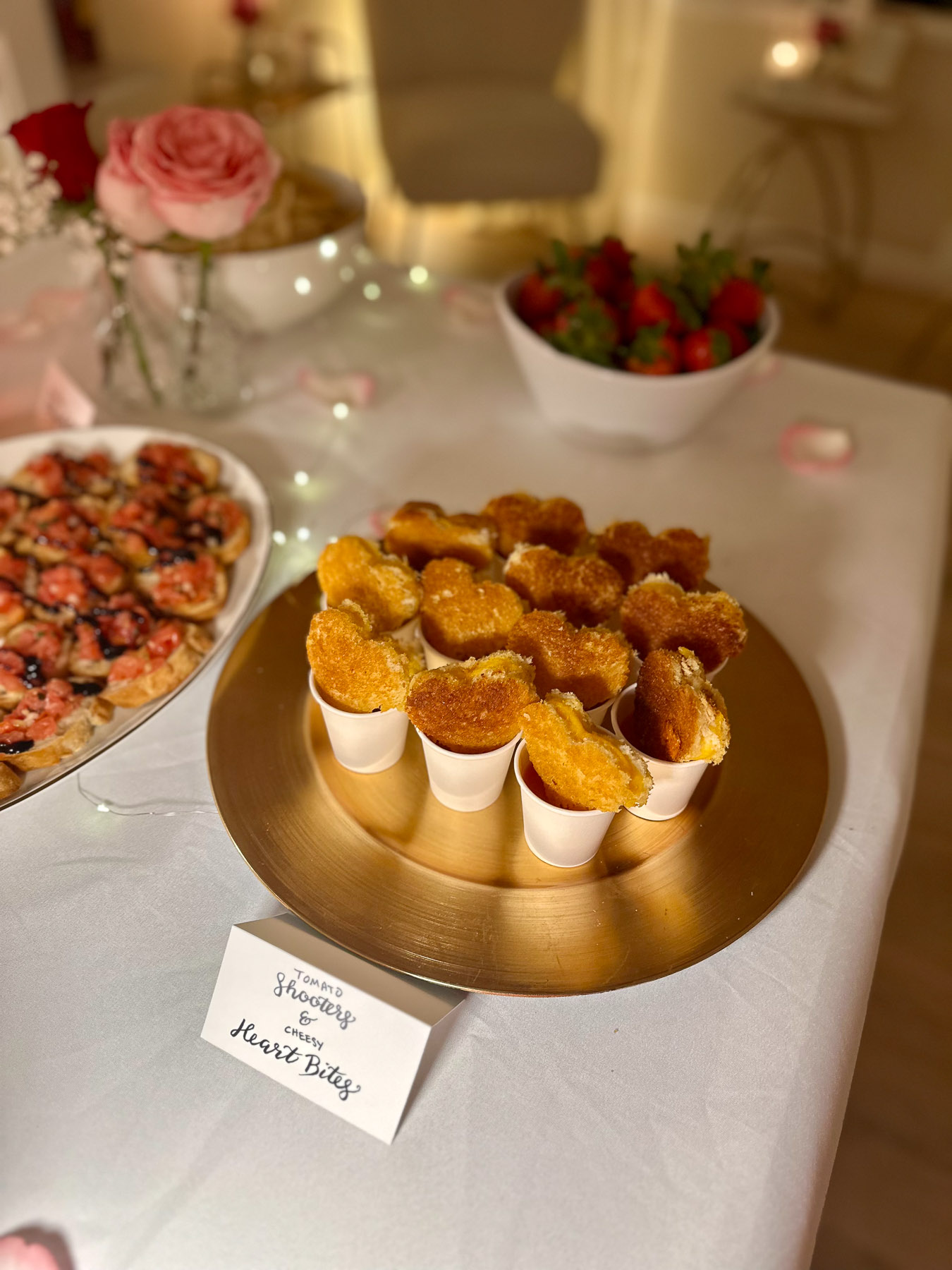 Mini grilled cheese hearts stacked in paper cups with tomato soup labeled “Tomato Shooters & Cheesy Heart Bites,” served on a gold tray with other appetizers nearby.