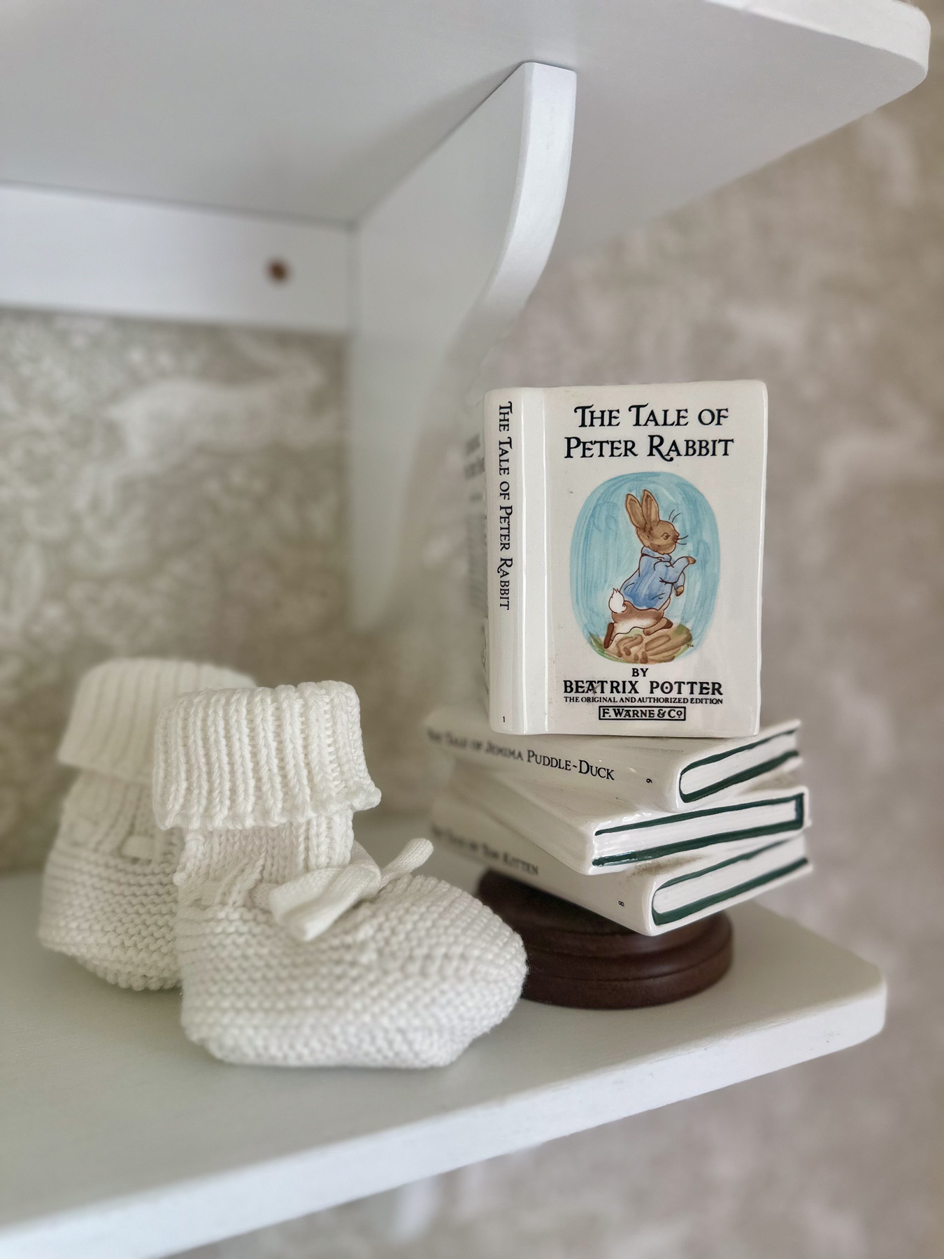 Ceramic Peter Rabbit book stack and knit baby booties styled on white nursery shelf decor.