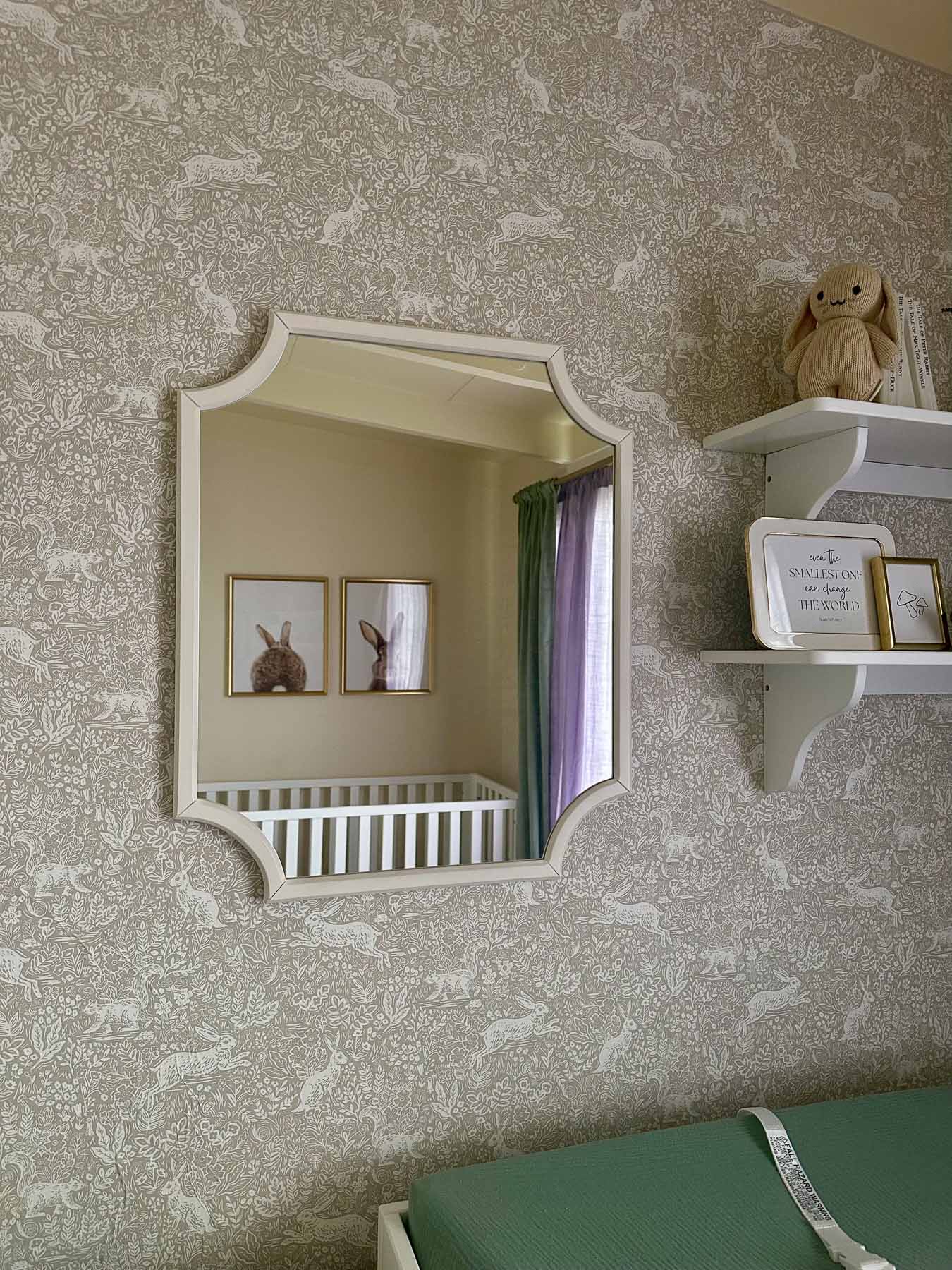 Scalloped wall mirror above a changing table with bunny-themed nursery wall decor and floating shelves.