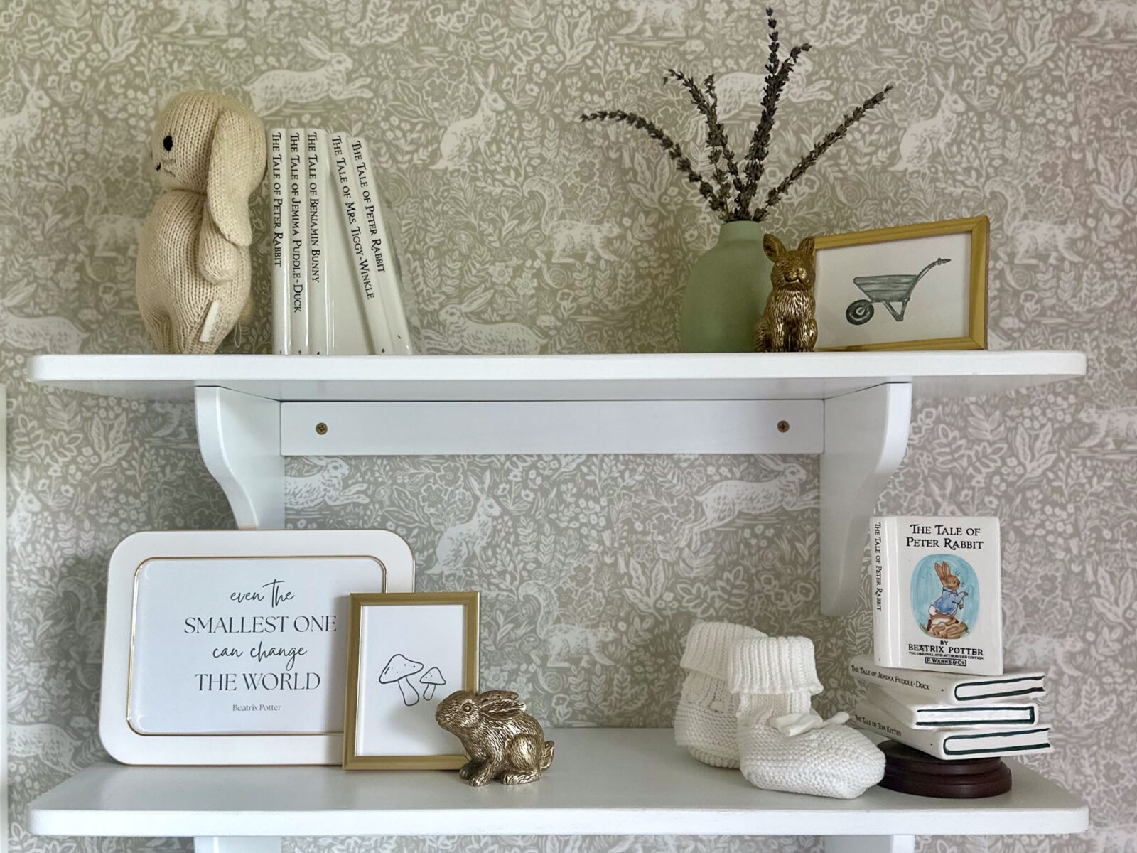 Neutral nursery shelf decor with bunny plush, children's books, baby booties, and framed art on white wall shelves.