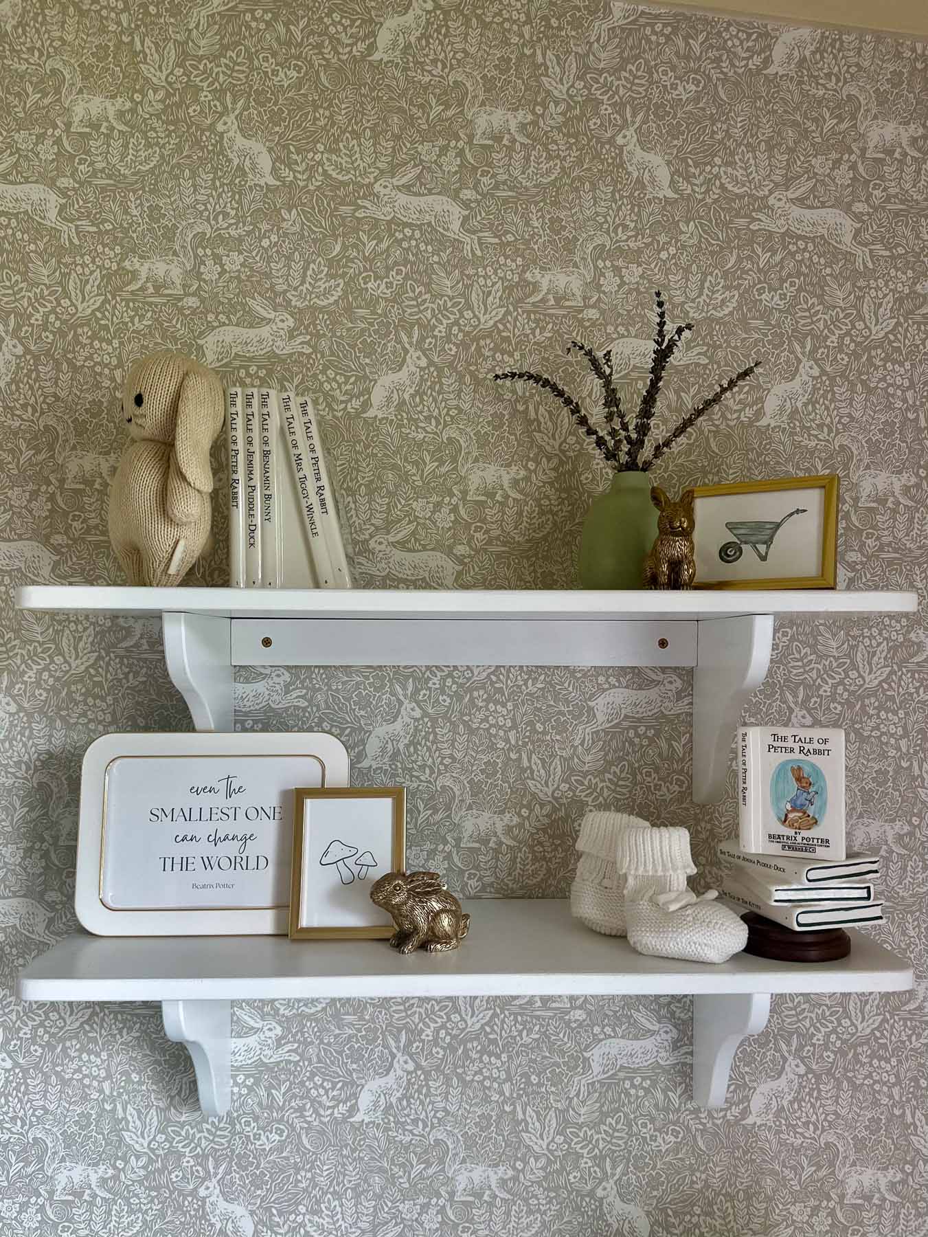 Neutral nursery shelf decor with knit bunny plush, children's books, framed prints, and ceramic books.