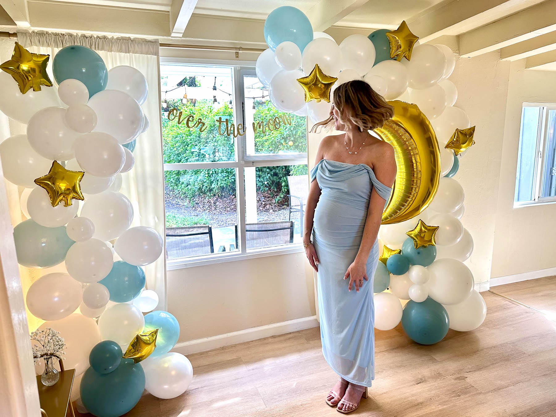 Pregnant woman in dusty blue dress standing between balloon arches with gold moon and star decor at an Over the Moon baby shower