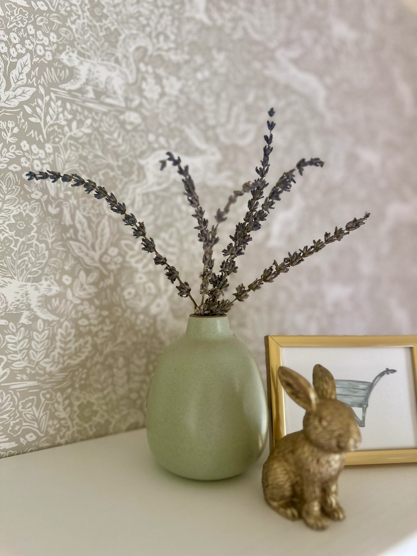 Green vase with dried stems and small brass bunny figurine beside framed wheelbarrow art on nursery shelf.