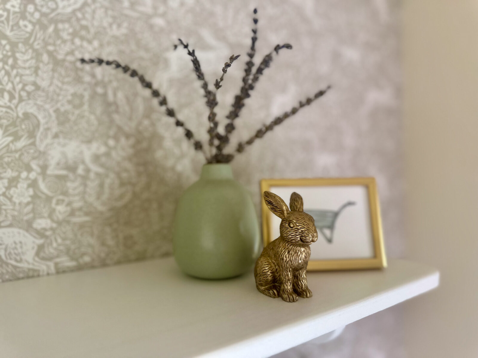 Green vase with dried stems and brass bunny figurine styled on nursery shelf decor.