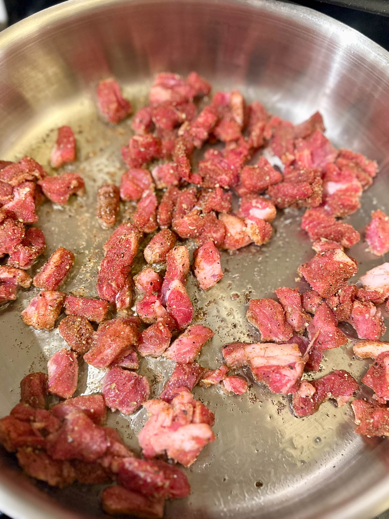 Raw seasoned ribeye pieces starting to sear in a stainless steel pan.