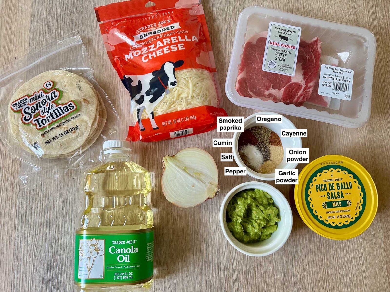 Ingredients for ribeye tacos including Trader Joe's Sonora style tortillas, ribeye steak, shredded mozzarella cheese, canola oil, half a sweet onion, pico de gallo, fresh guacamole, and a ramekin of spices (paprika, cumin, oregano, garlic powder, onion powder).