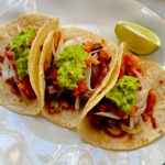 Three ribeye steak tacos on a white plate, featuring gold-brown cheese-crusted flour tortillas topped with grilled steak, fresh guacamole, pico de gallo, and sautéed onions, served with a lime wedge.
