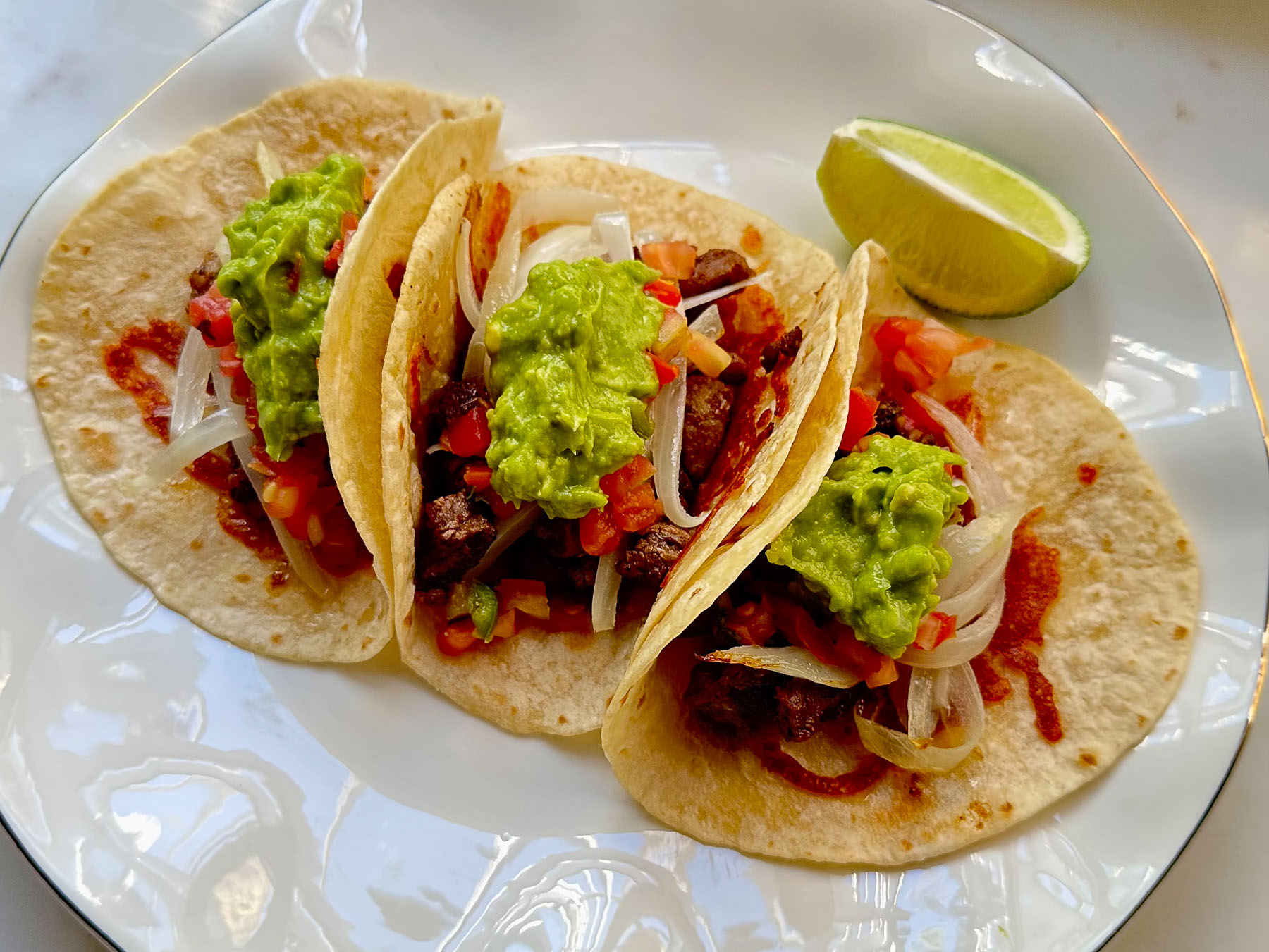 Three ribeye steak tacos on a white plate, featuring gold-brown cheese-crusted flour tortillas topped with grilled steak, fresh guacamole, pico de gallo, and sautéed onions, served with a lime wedge.