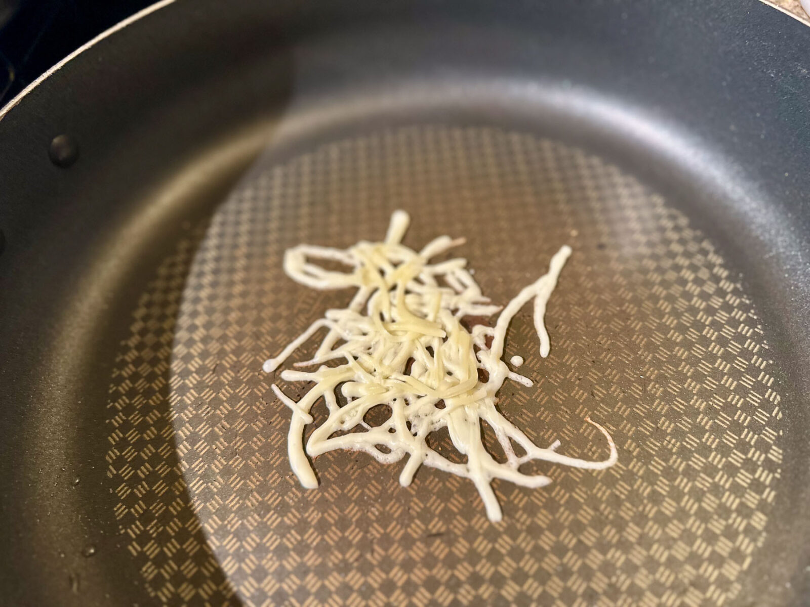 Shredded mozzarella cheese melting and bubbling in a circular shape in a nonstick skillet.
