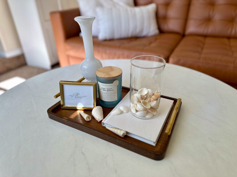 A wooden coffee table tray styled with coastal summer decor, including loose seashells, a candle, and a framed wave illustration on a white marble table.