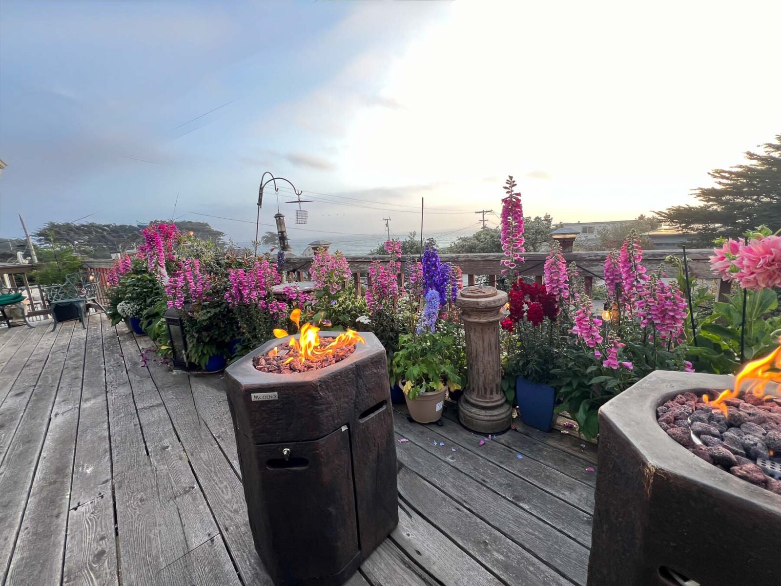 Two lit hexagonal fire pits on a wooden deck lined with potted pink and purple flowers, overlooking an ocean view at sunset.