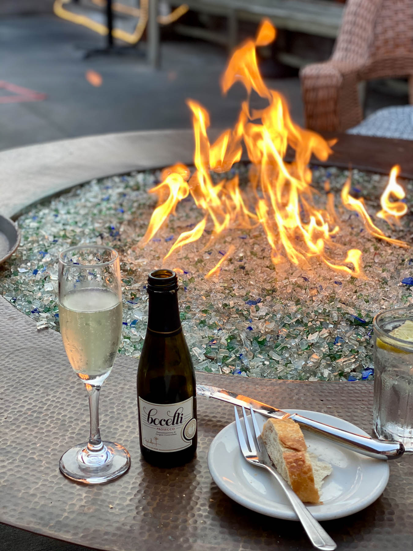 Close-up of a flaming fire pit filled with glass rocks, featuring a glass of champagne and a small appetizer resting on the copper rim.