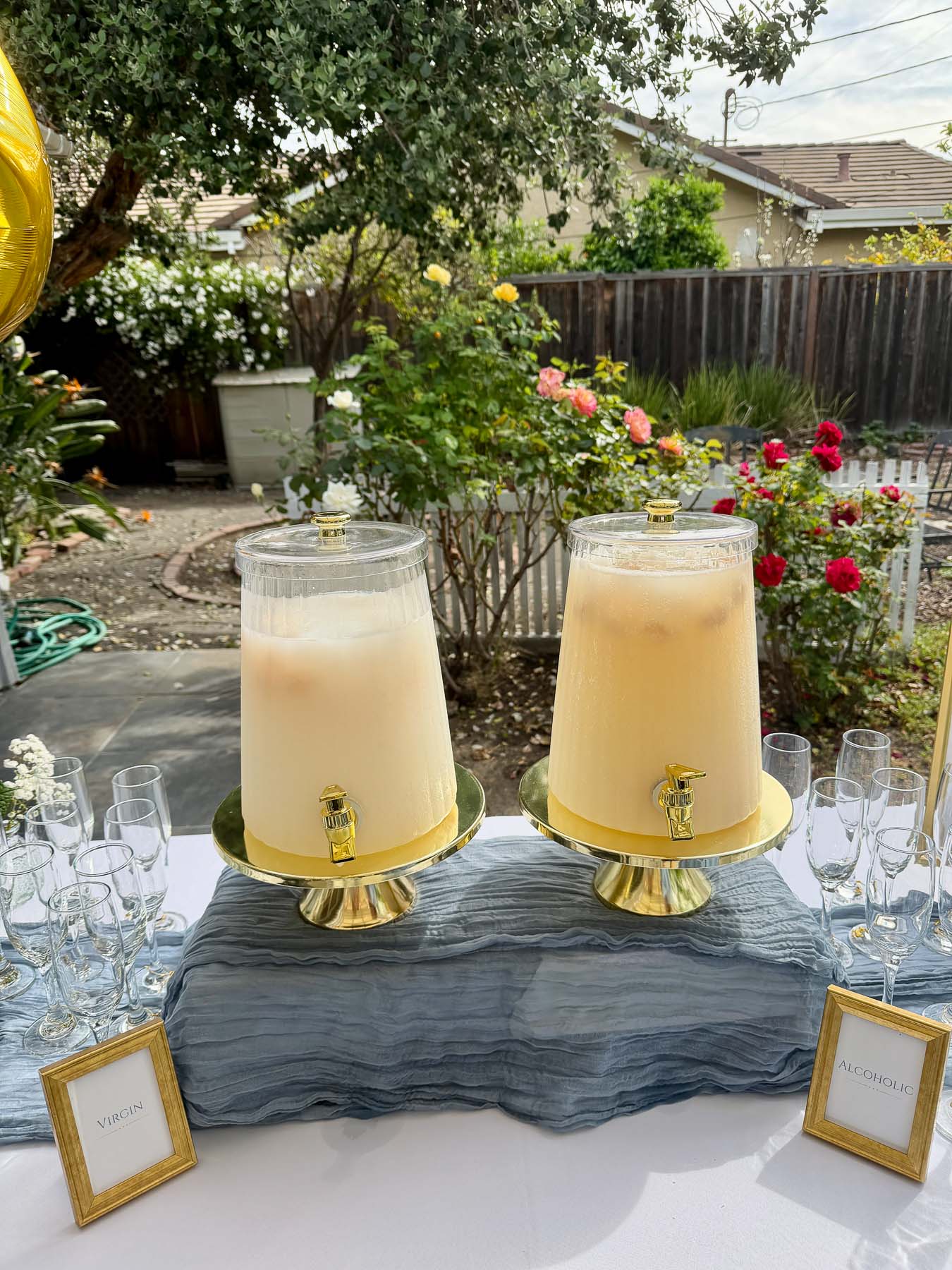Two glass drink dispensers on gold stands over a blue runner, an elegant beverage station backyard setup.