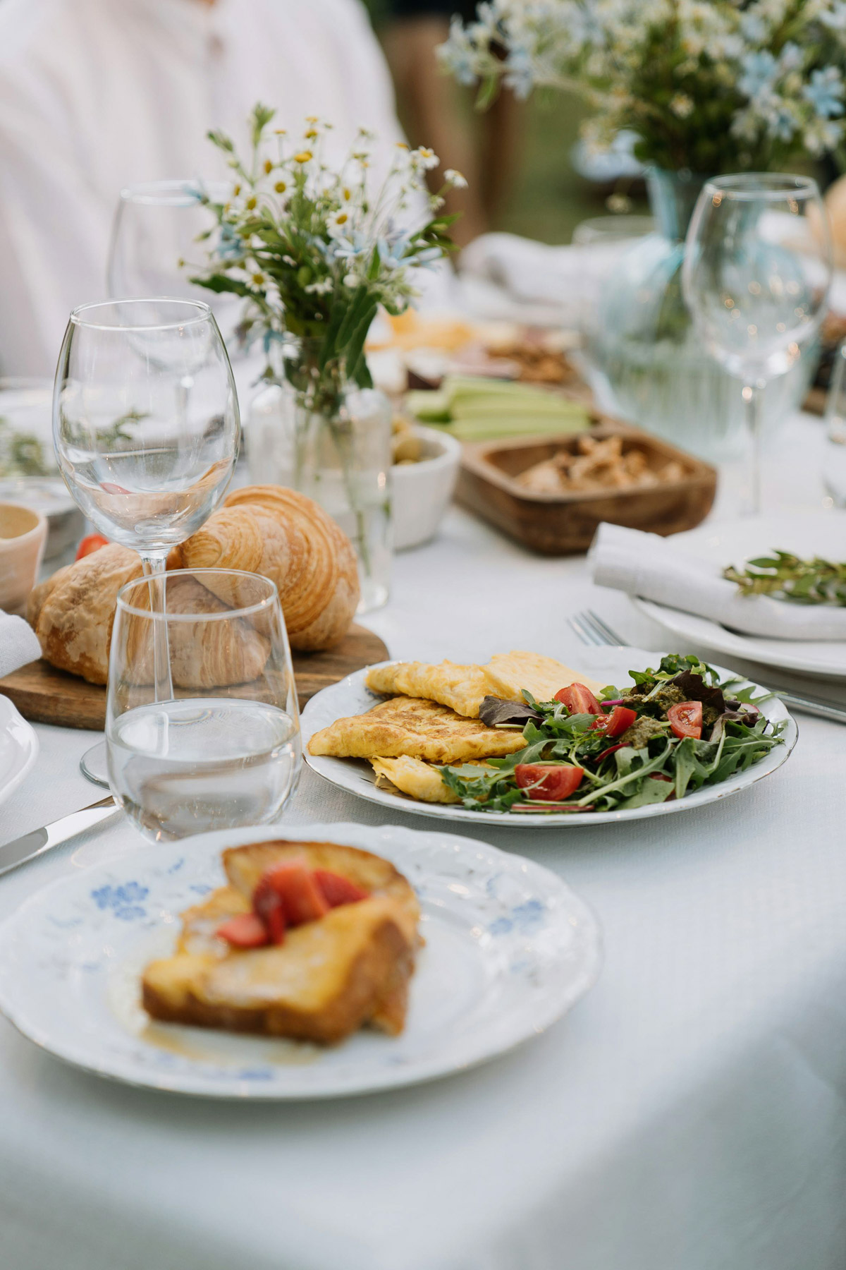 An elegantly set outdoor dining table featuring plates of food, a wooden bowl of croissants, wine glasses, and chamomile floral centerpieces for a backyard brunch.