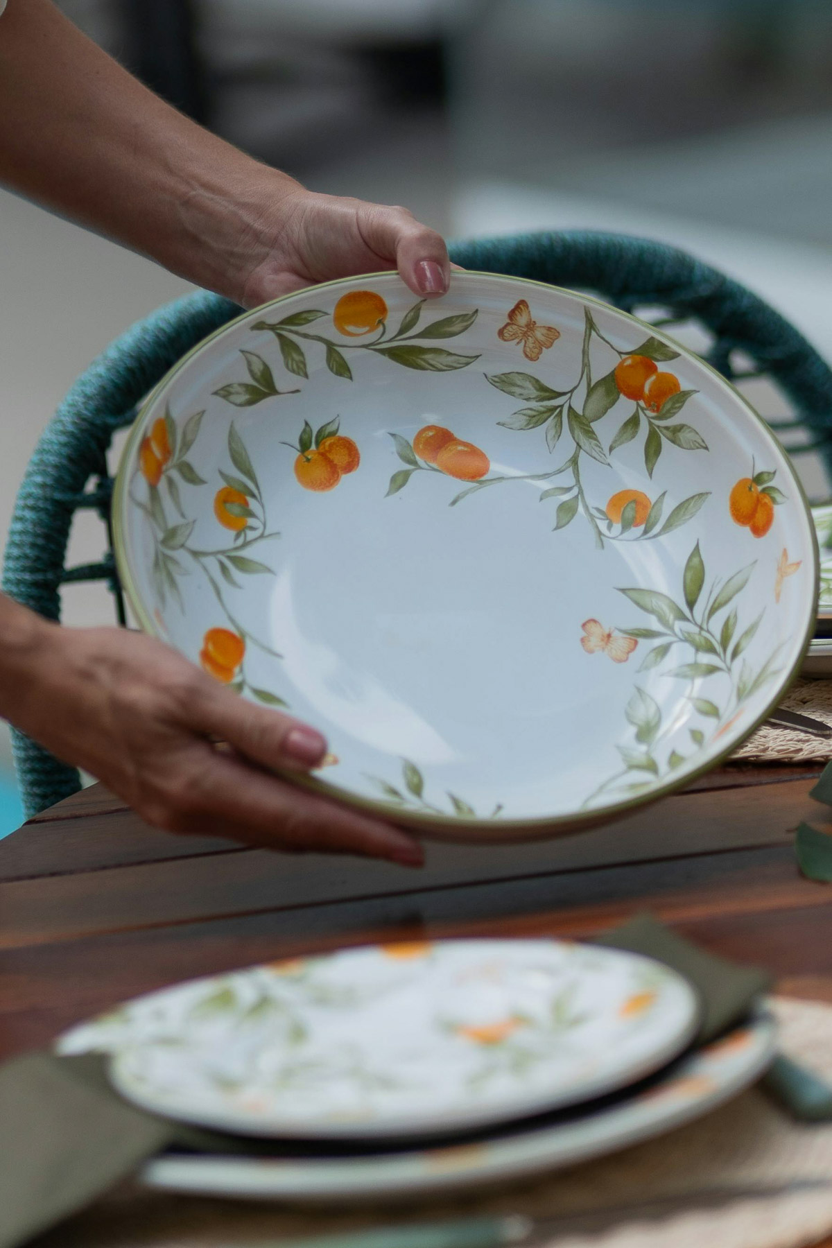 Hands holding a white ceramic ourdoor serving bowl decorated with painted oranges.