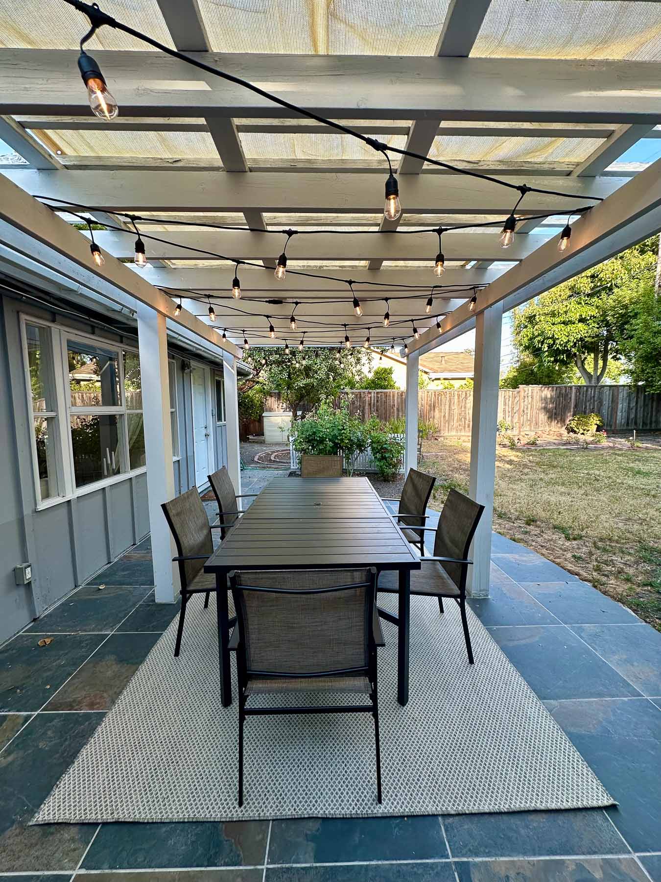 A spacious dark outdoor dining table and chairs on a rug under a white pergola, a classic foundation for backyard entertaining ideas.