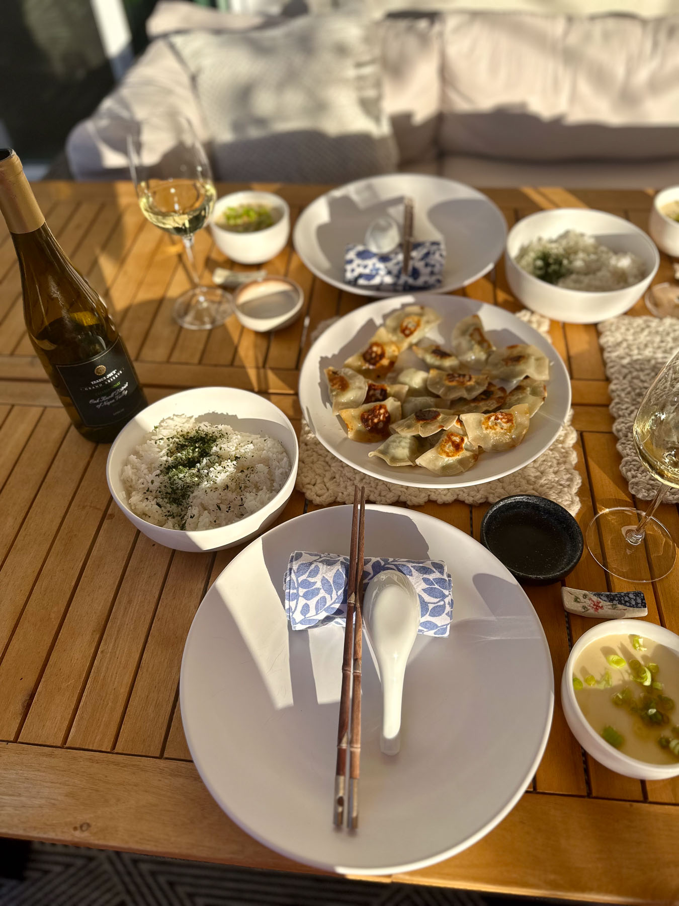 A slatted wooden outdoor table beautifully set with plates of pan-fried dumplings, bowls of rice and soup, chopsticks, and white wine in the warm golden hour sun.