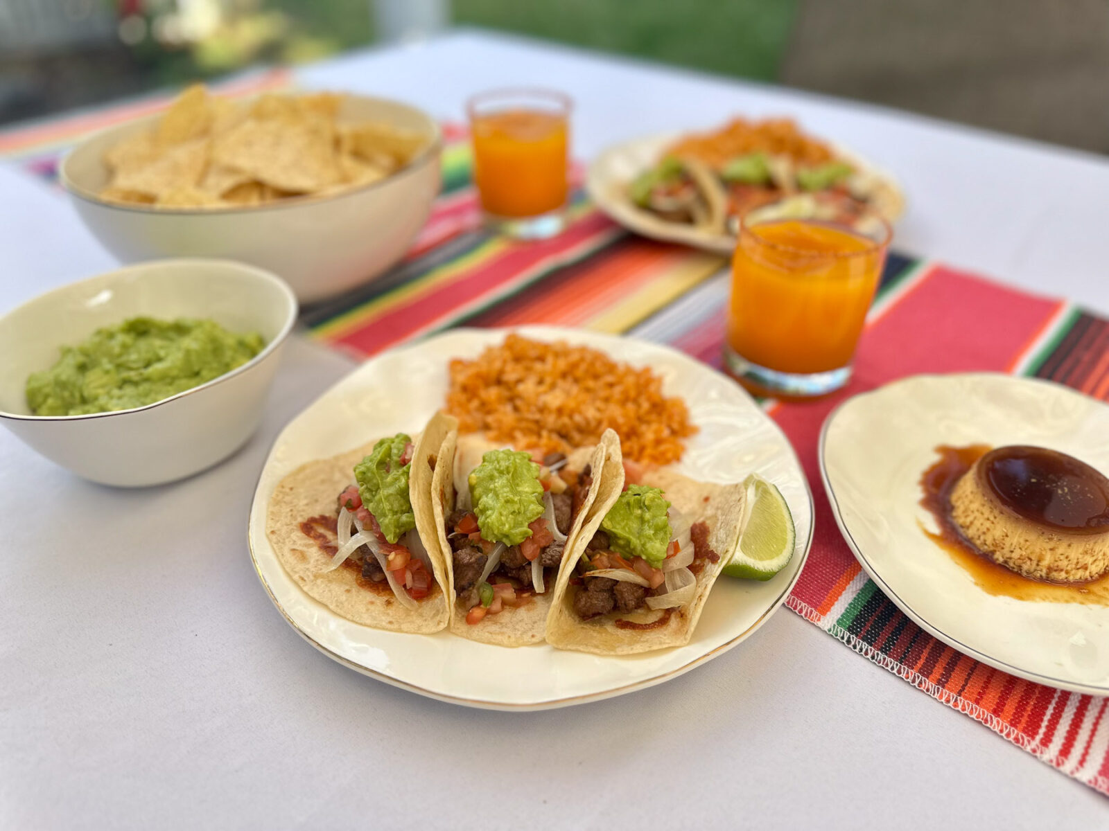 A festive outdoor table setting featuring steak tacos, guacamole, rice, and flan served on white and gold dishes and a colorful striped table runner.